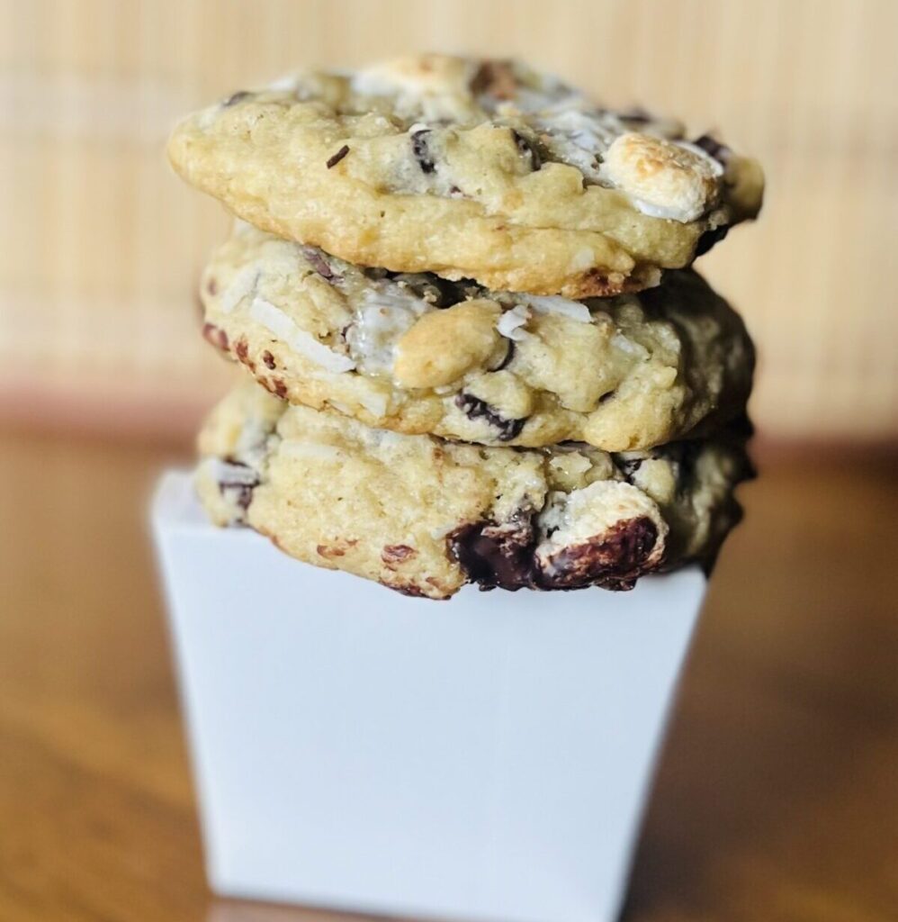 Stack of three chocolate chip, coconut, marshmallow cookies on top of takeout container on a table